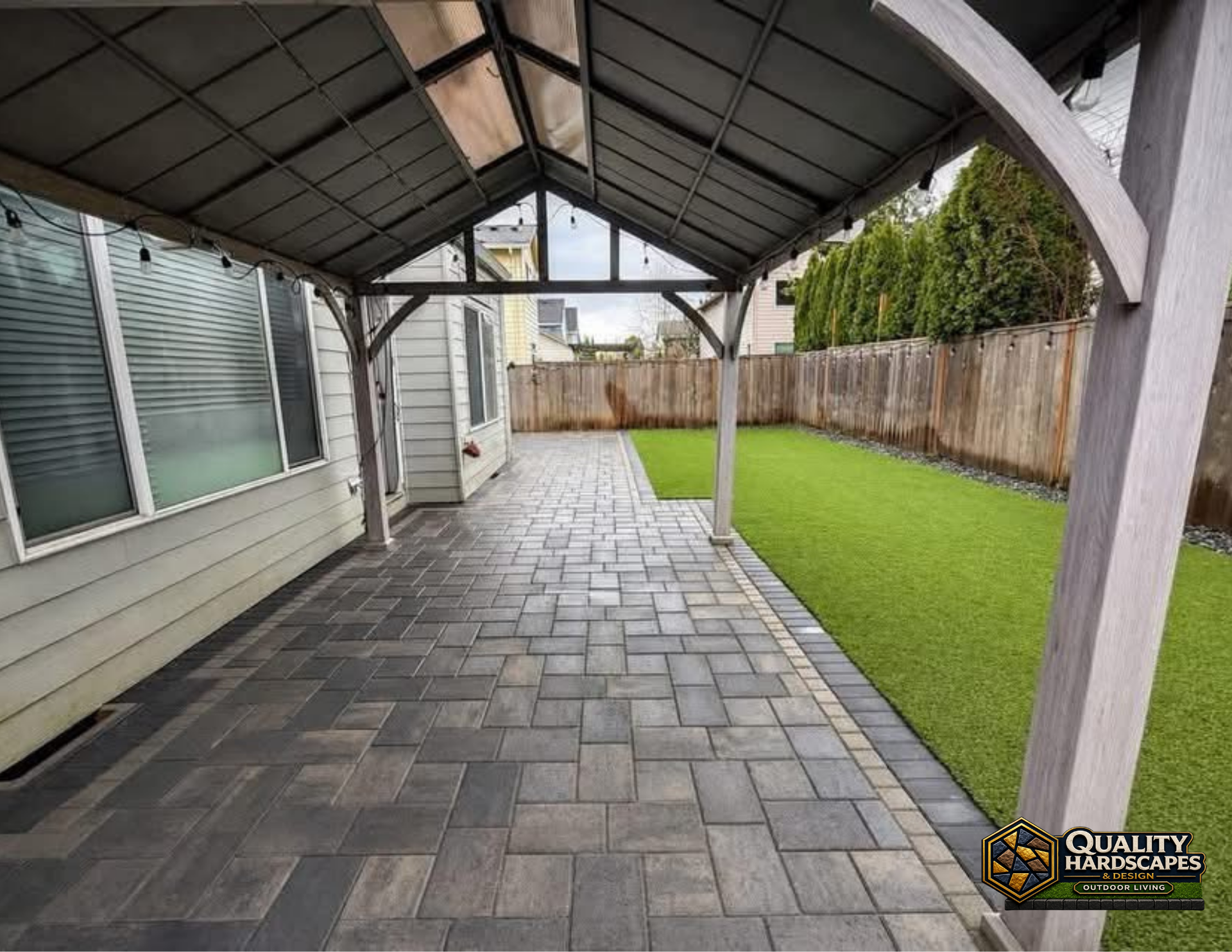 Covered patio with gray pavers, white siding, and a green artificial grass strip along the side yard