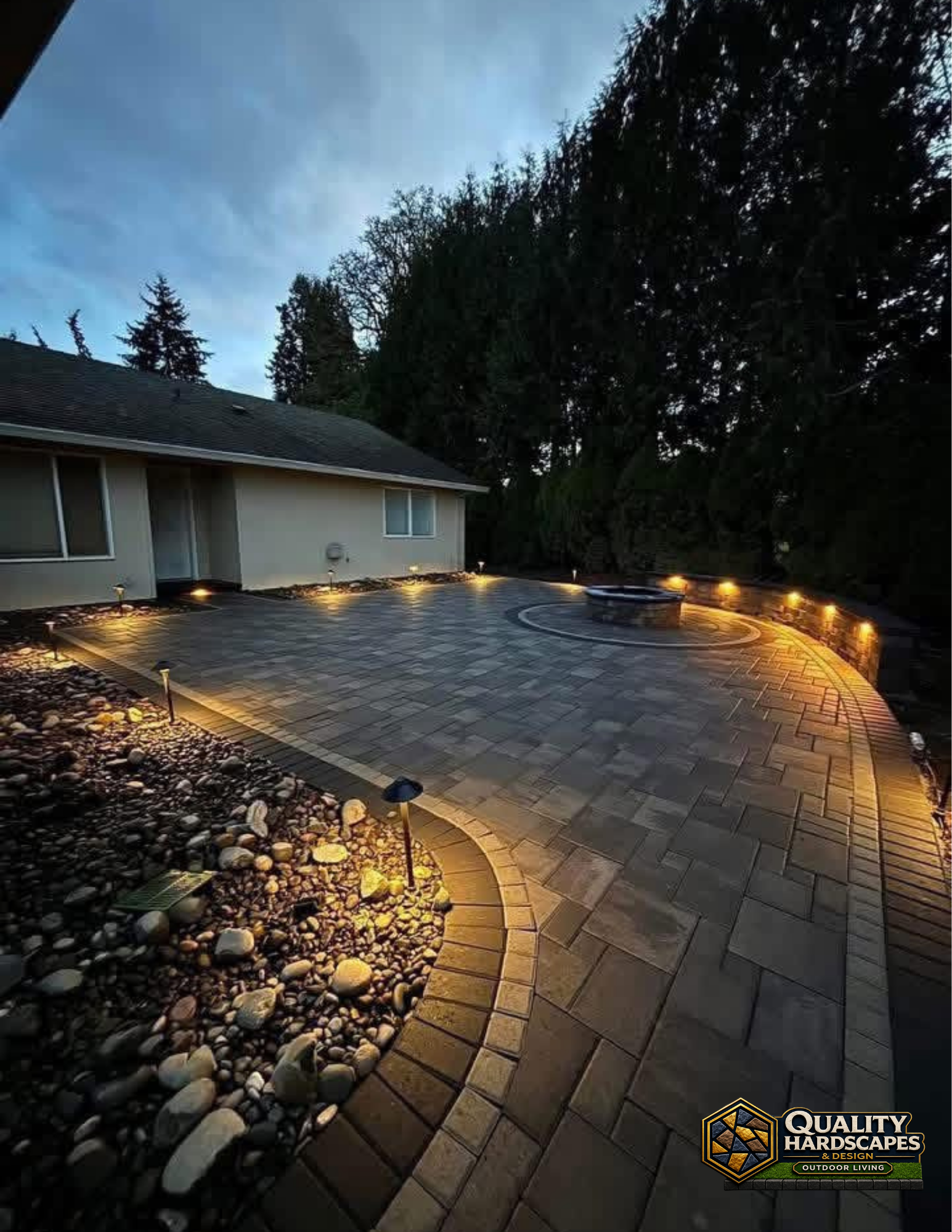 Curved stone patio with warm path lights beside a house at dusk, bordered by trees and rock landscaping