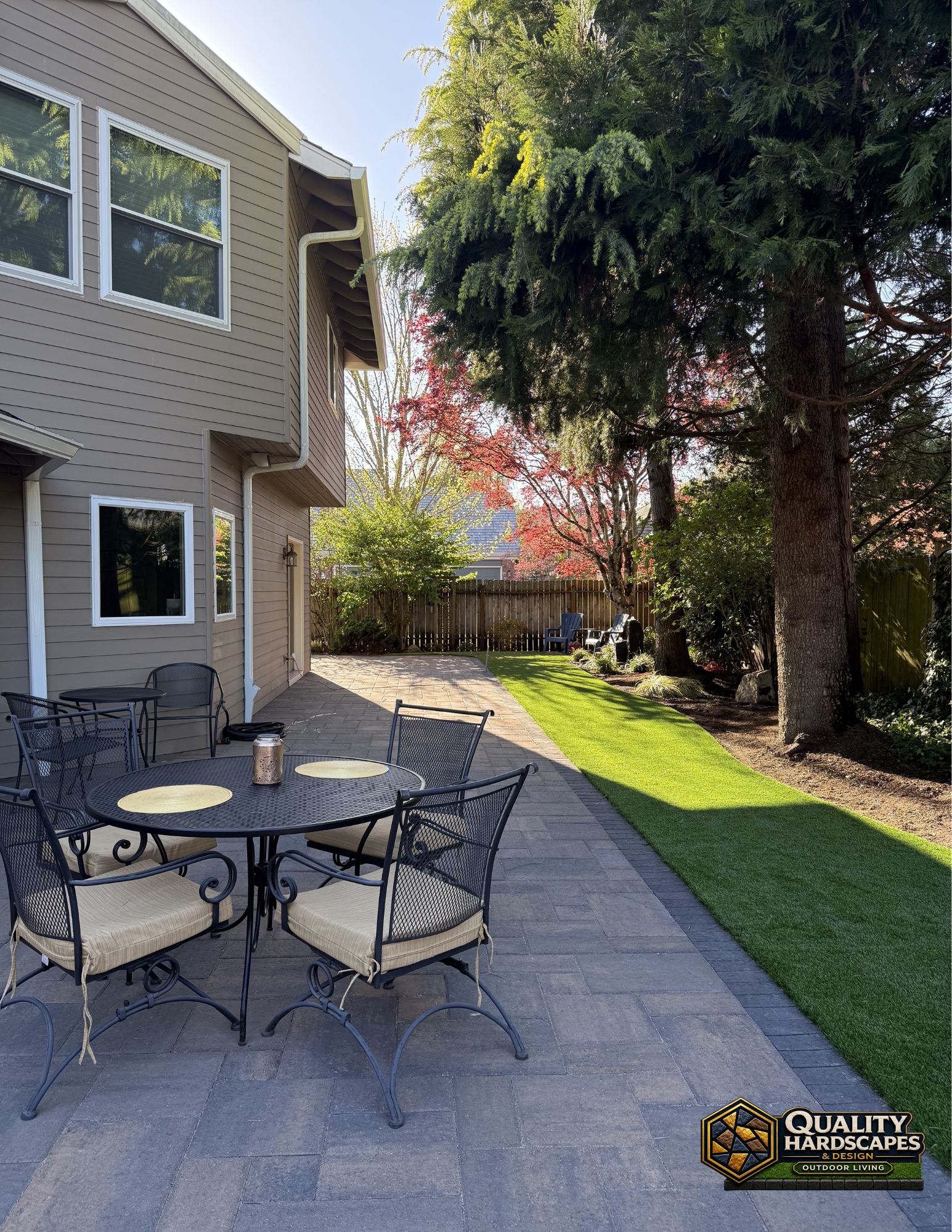 Patio with outdoor table and chairs beside a house, overlooking a sunny backyard and trees.