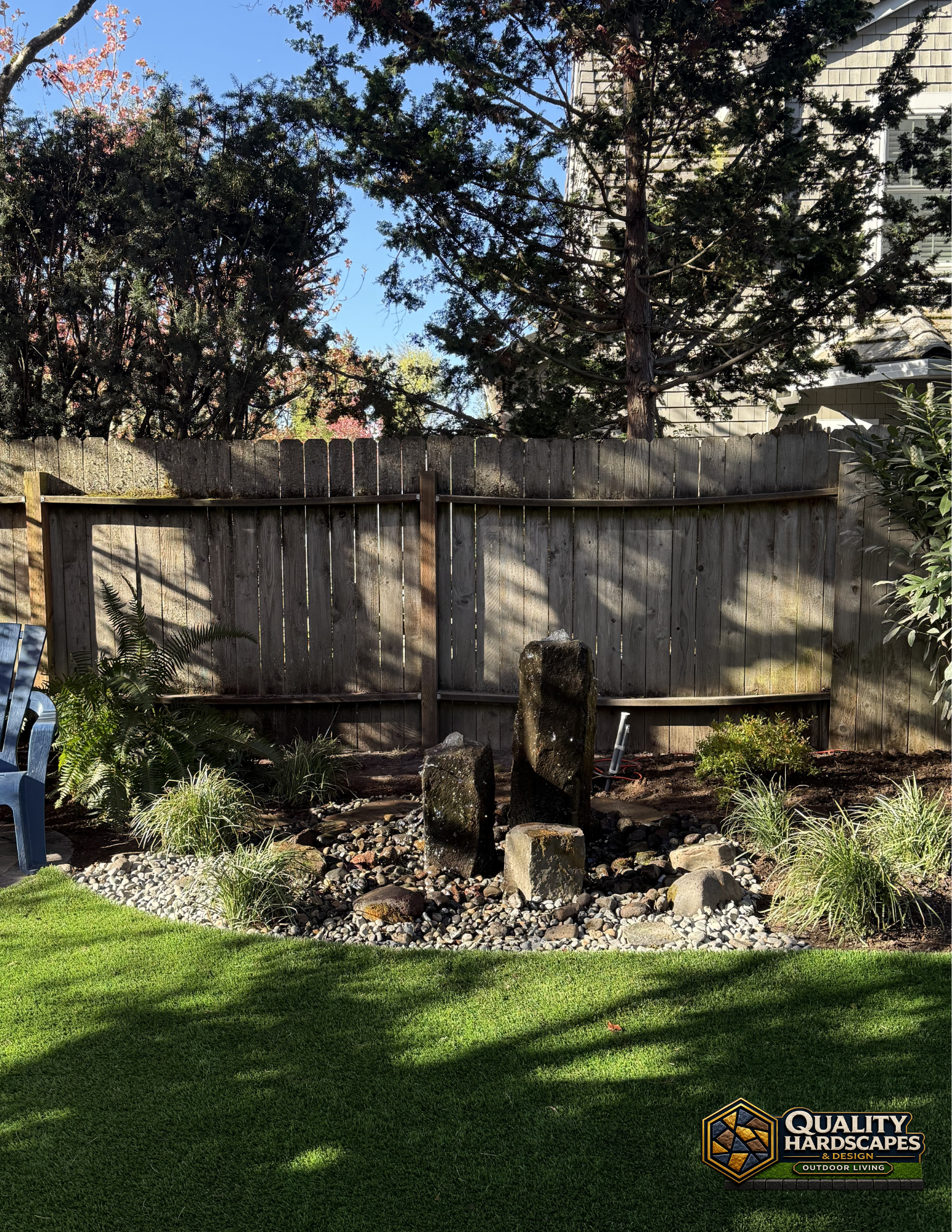 Backyard garden with a small stone fountain, wooden fence, and green lawn in sunlight