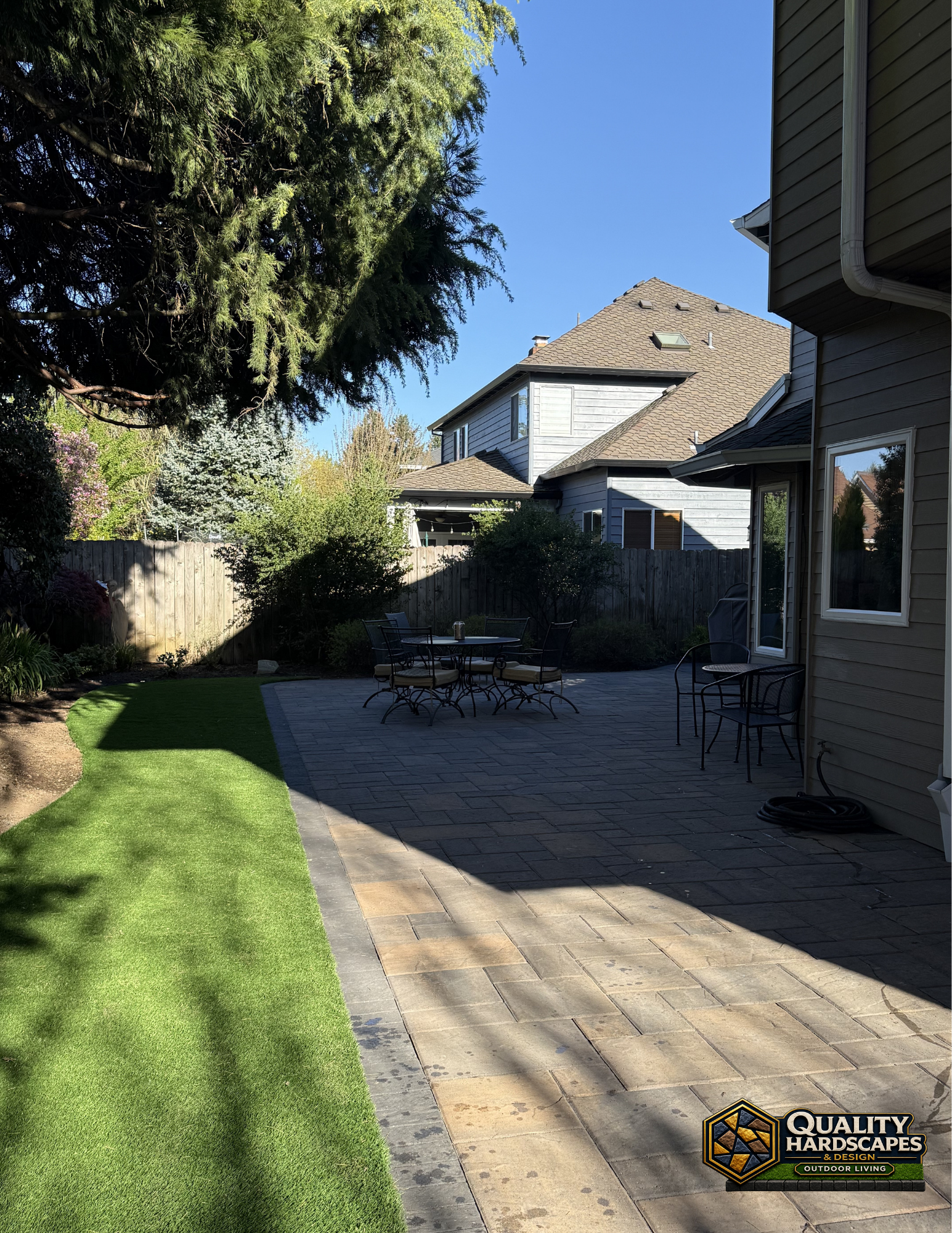 Backyard patio with stone pavers, green lawn, trees, and a house under a clear blue sky