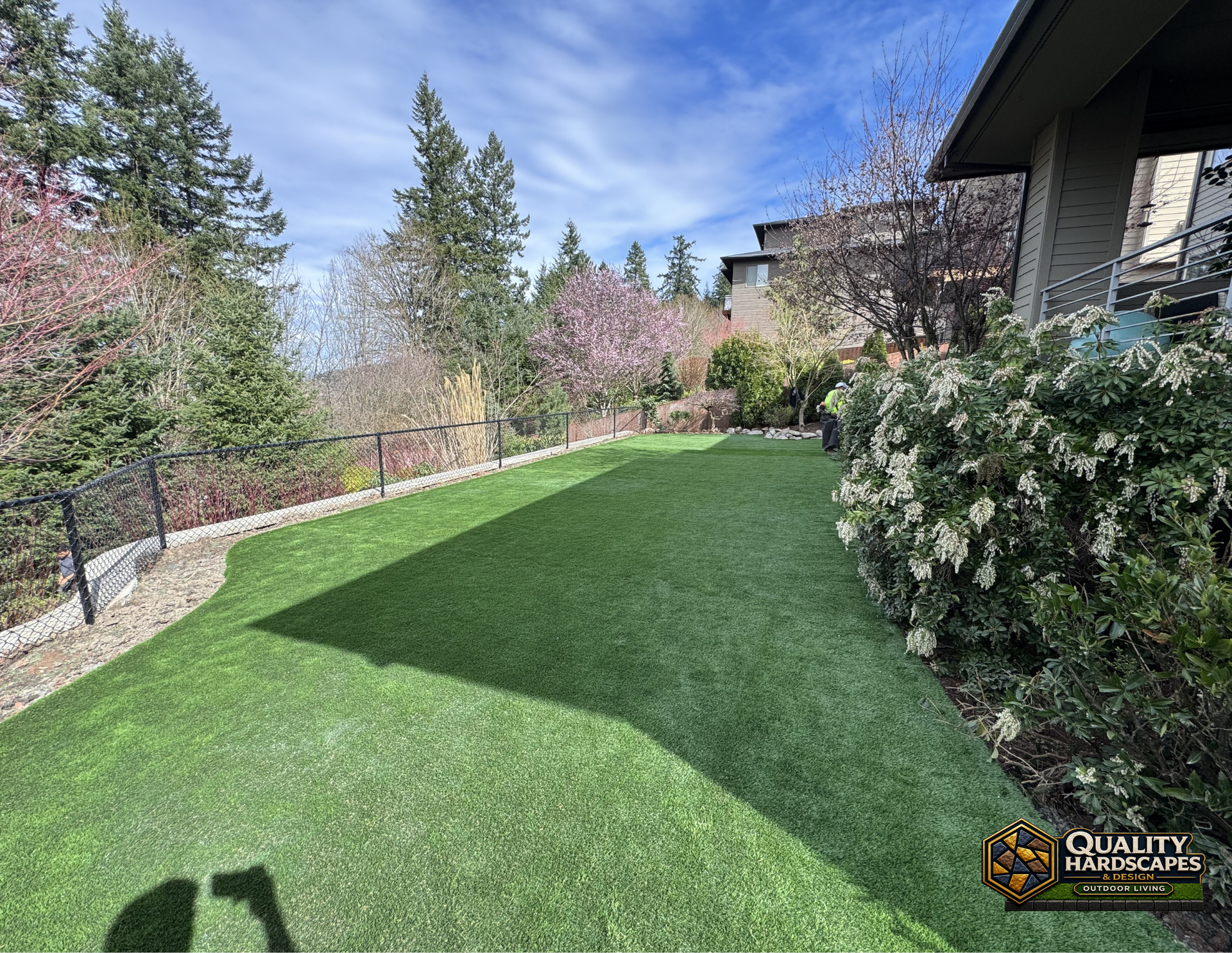 Landscaped backyard with lush green lawn, blooming trees, and a patio beside a house.
