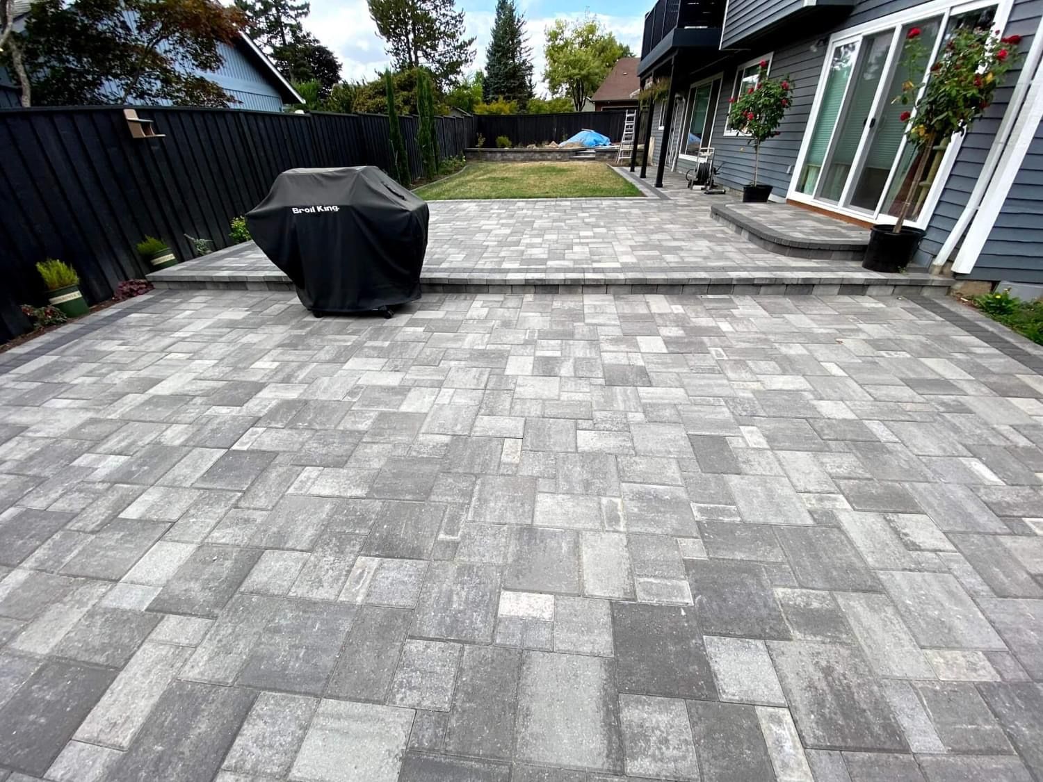 Paved patio with dark grill cover; house in background; black fence on left; lawn beyond.