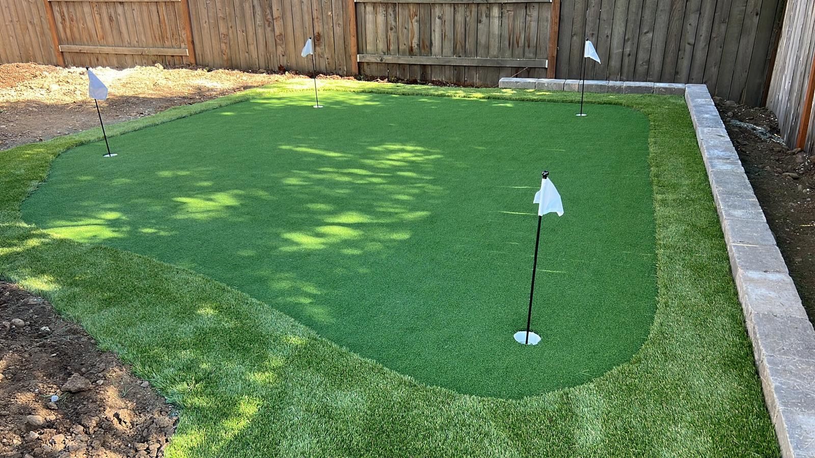 Backyard putting green with multiple holes and flags, surrounded by grass and a retaining wall.