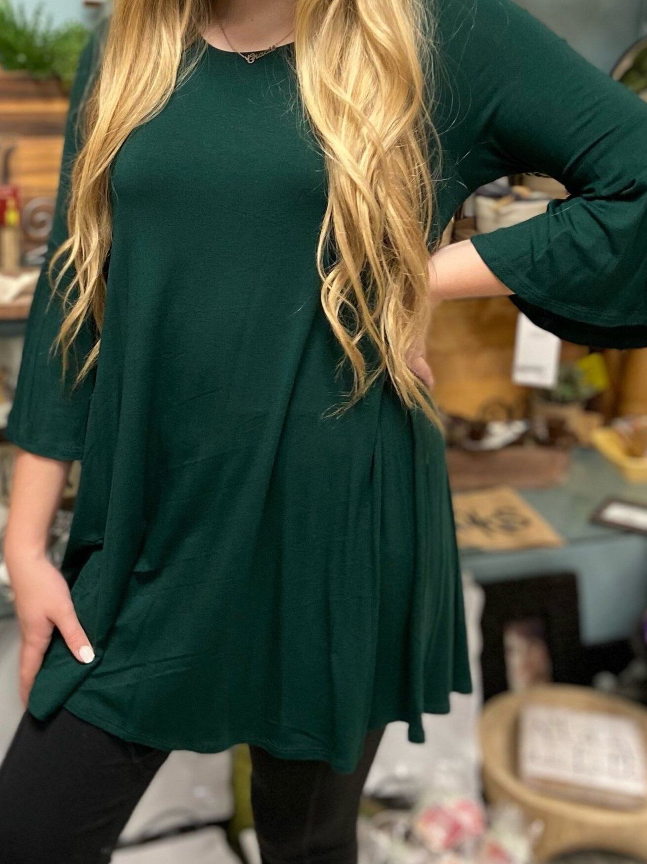 A woman wearing a green tunic and black pants is standing in a room.