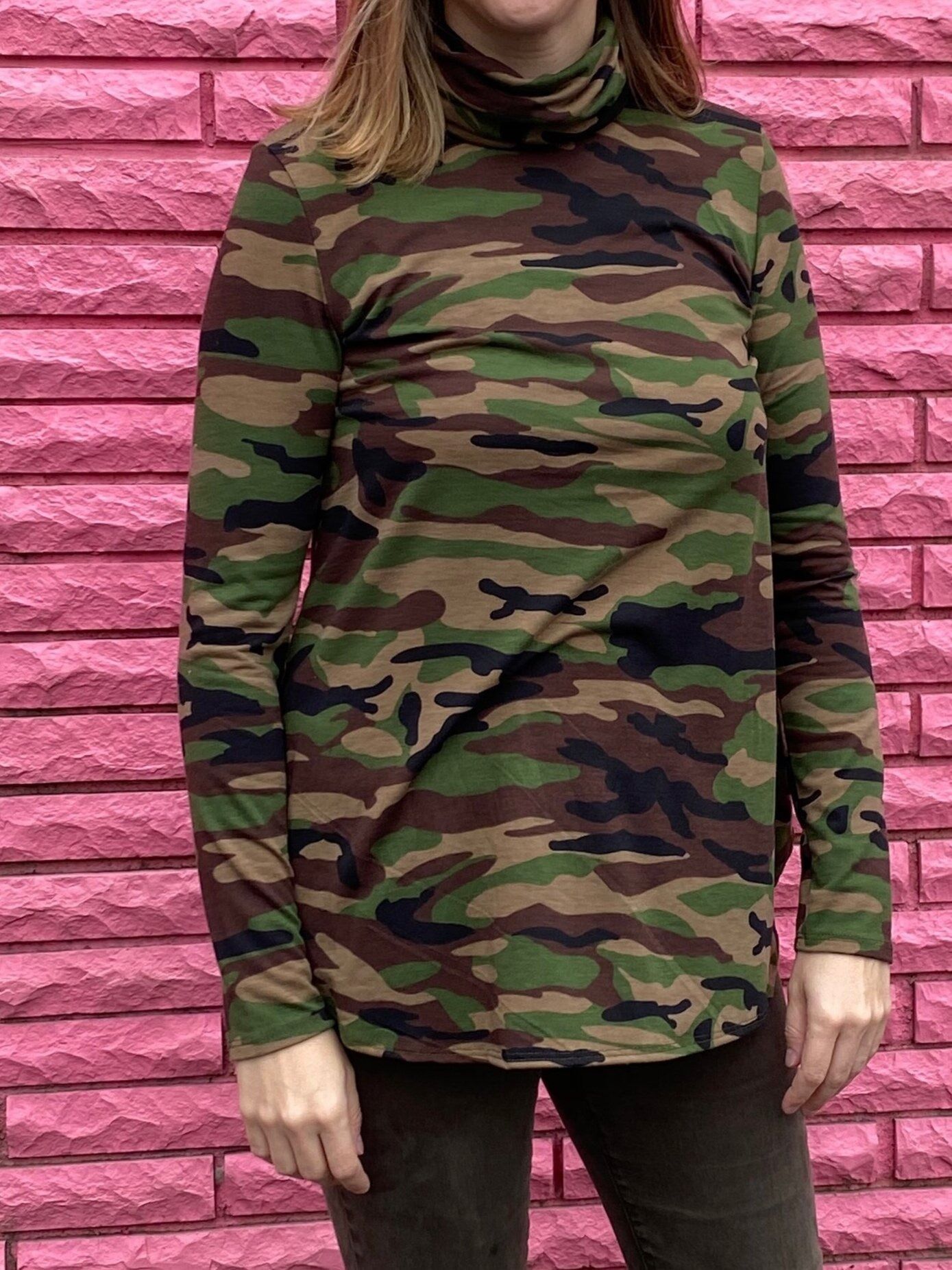 A woman wearing a camouflage turtleneck sweater is standing in front of a pink brick wall.
