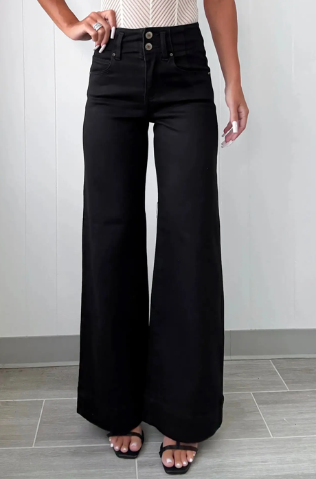 Woman wearing black high-waisted wide-leg jeans, standing in front of a white wall.