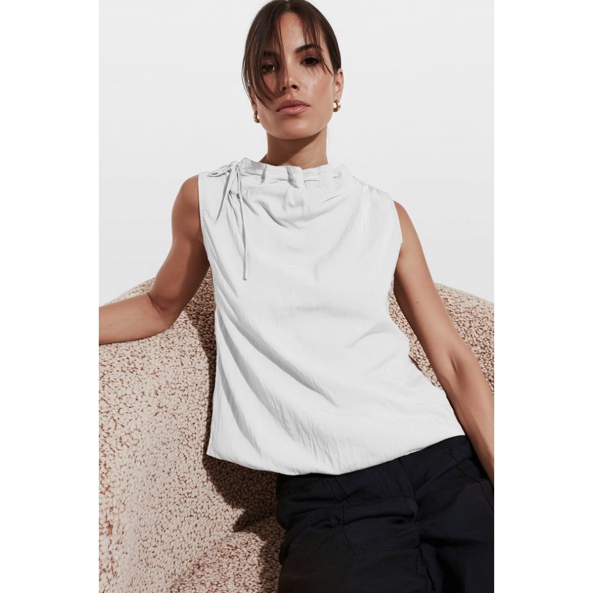 Woman in white sleeveless top and black pants. Leaning against a beige chair.