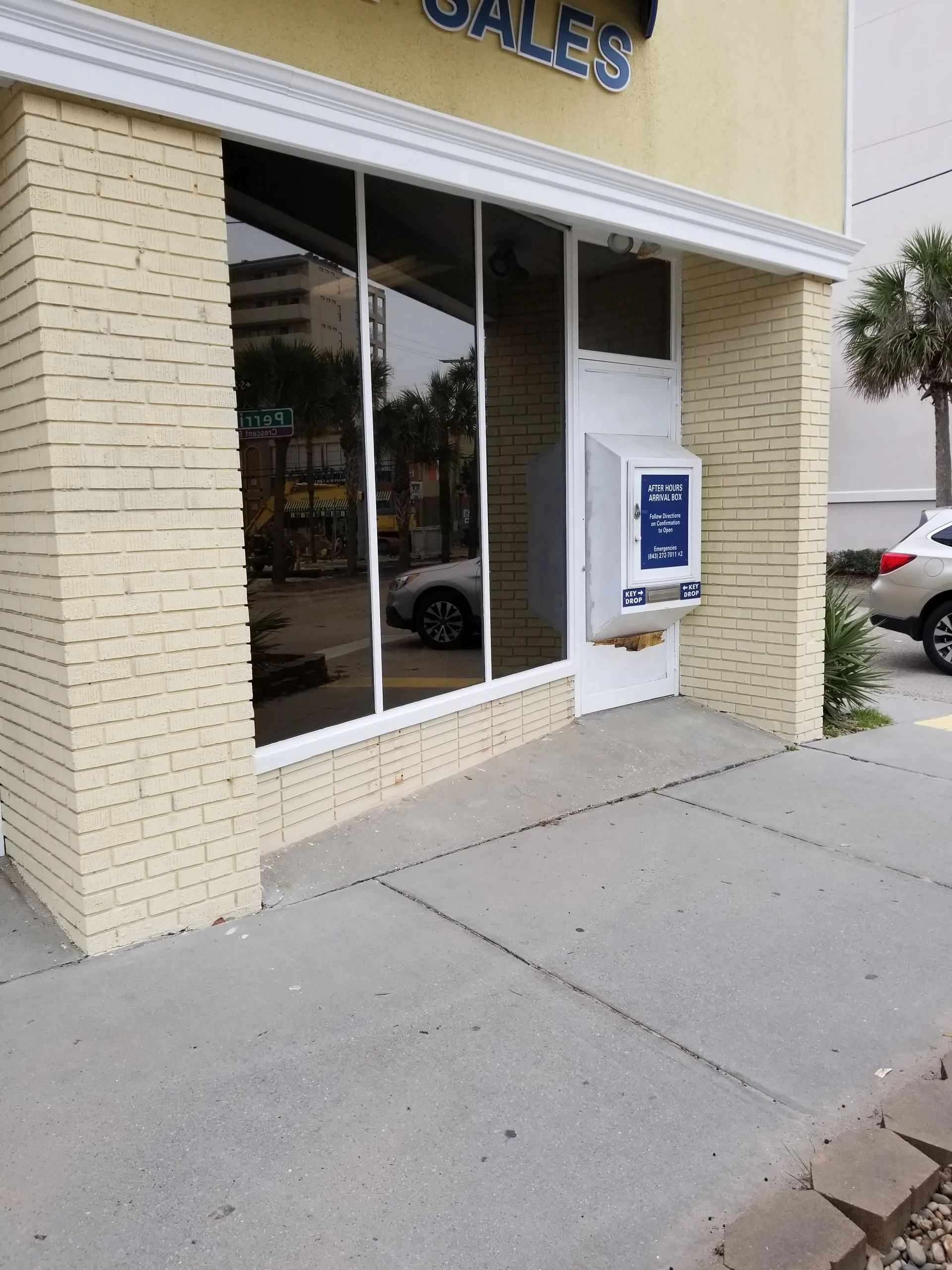 Exterior of a store with large windows and a white brick facade. A car is visible in the reflection.