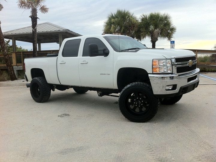 White lifted Chevrolet pickup truck on a sandy surface, black wheels, palm trees in the background.