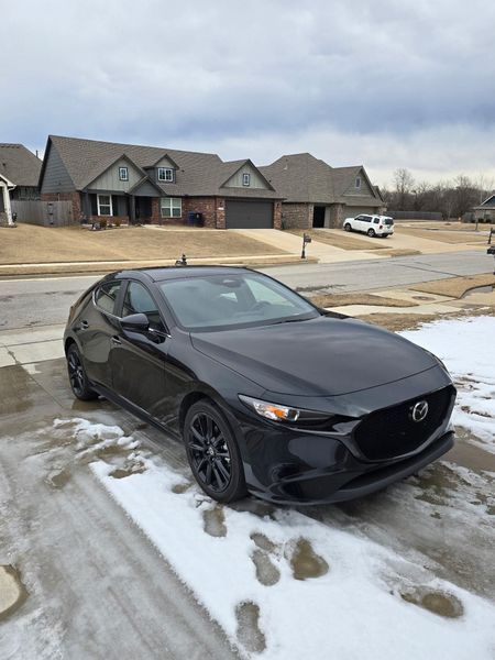 Black Mazda 3 hatchback parked on a snow-covered driveway in front of suburban houses. Cloudy sky.