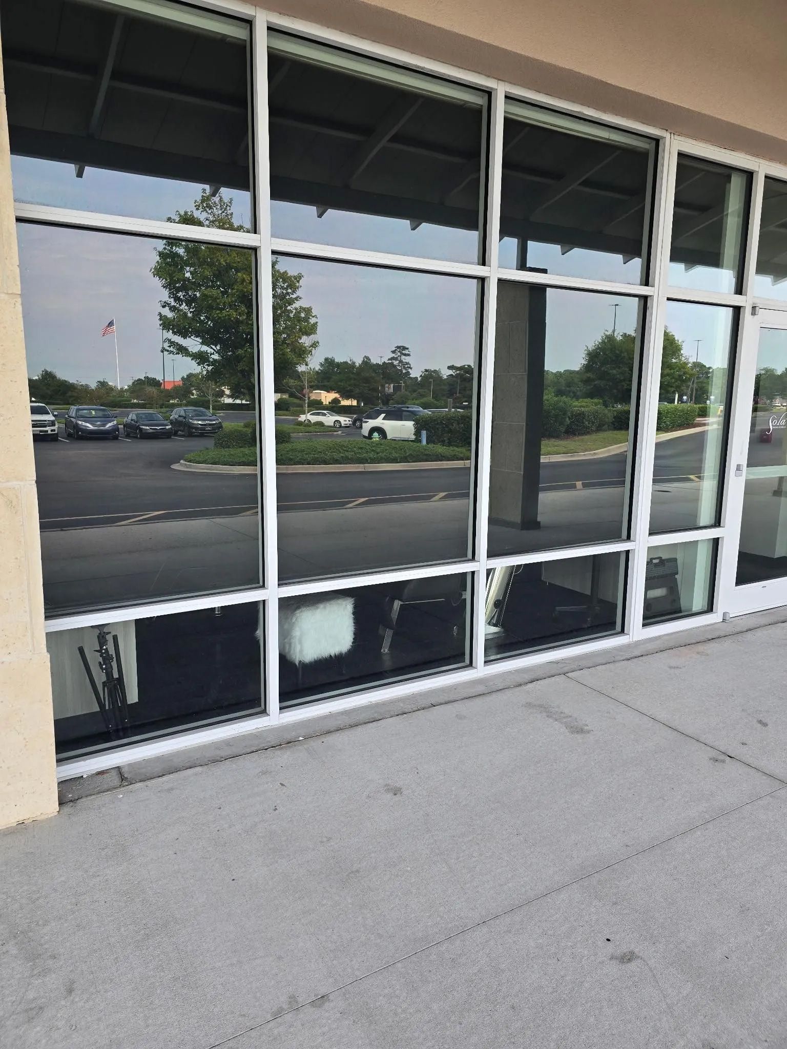 Large storefront windows reflecting a parking lot and sky, framed by white. Concrete sidewalk.