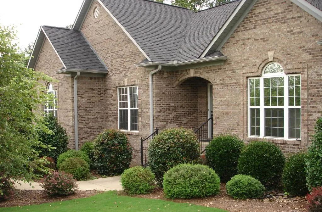 Brick house with dark roof and white-framed windows, surrounded by green shrubs and a small front yard.
