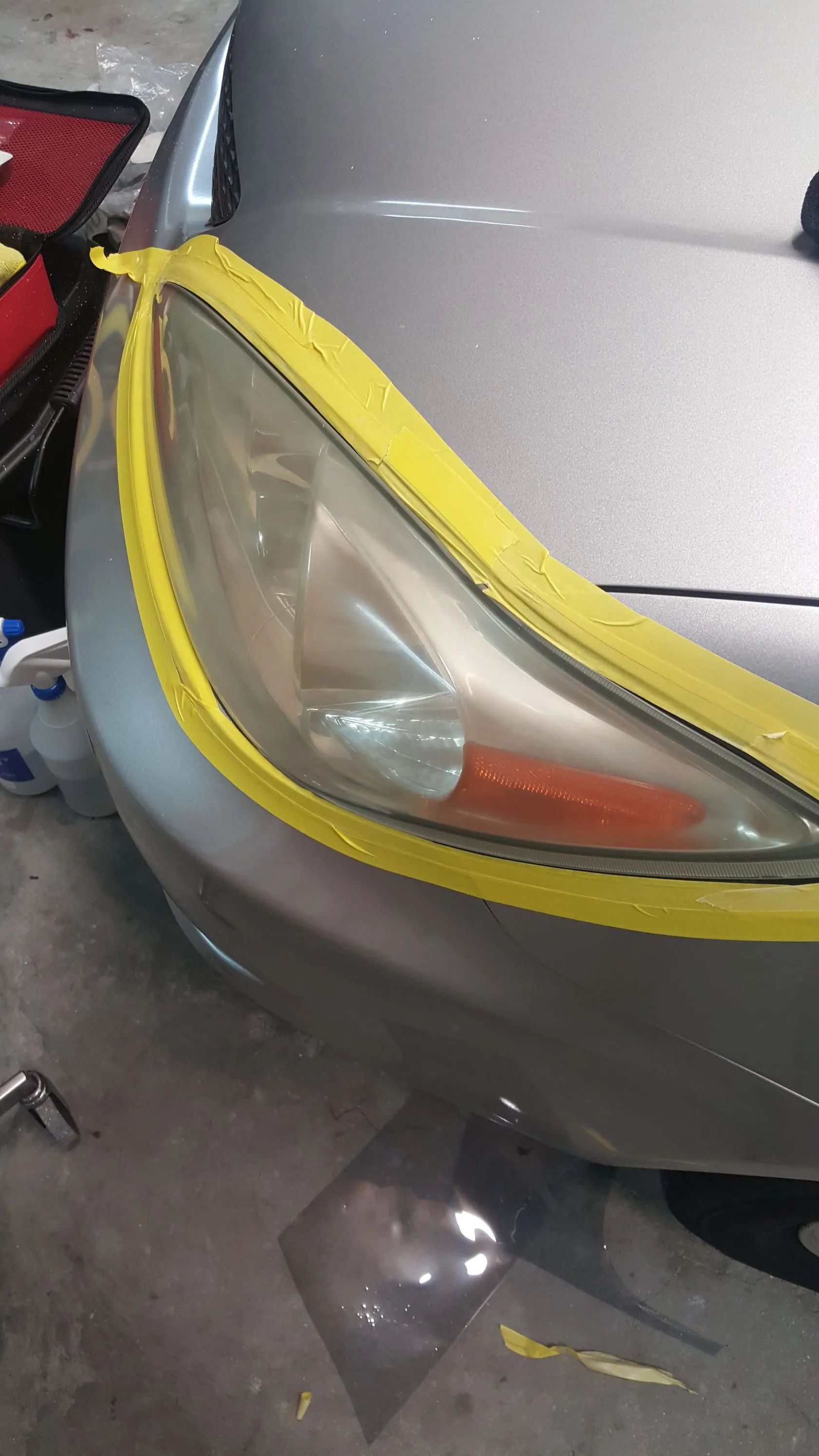A car headlight taped with yellow masking tape is being restored; gray car body.