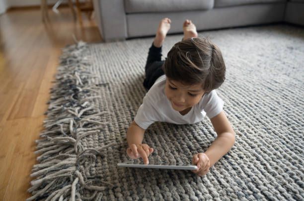 Child laying on a rug, using a tablet. They wear a white shirt and black pants.