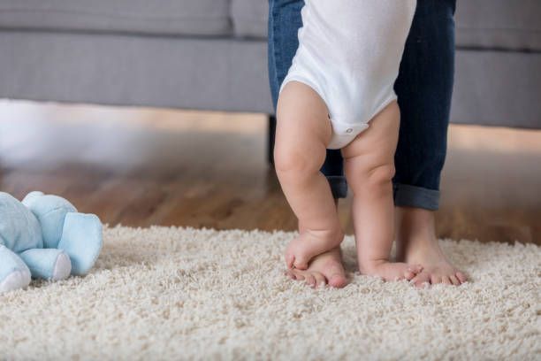 Baby learning to walk, held by an adult's legs. The baby is wearing a white onesie, standing on a fluffy rug.