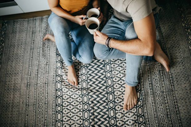 Couple sitting on a rug, holding coffee mugs, legs crossed, bare feet.