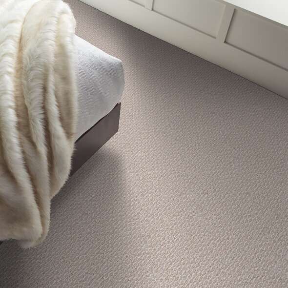 Beige carpeted floor next to a bed with a white comforter and built-in white cabinets.