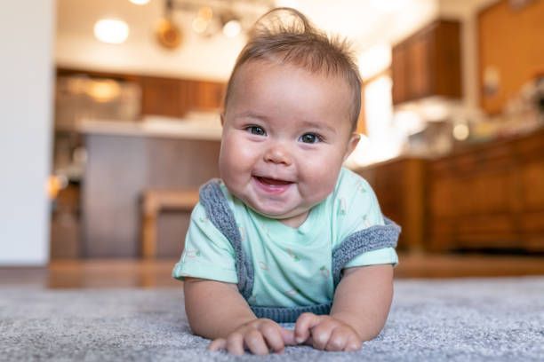 Happy baby on a gray carpet, smiling at the camera in a kitchen.