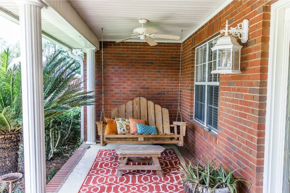 Covered porch with brick wall, swing, colorful cushions, rug, and potted plants.