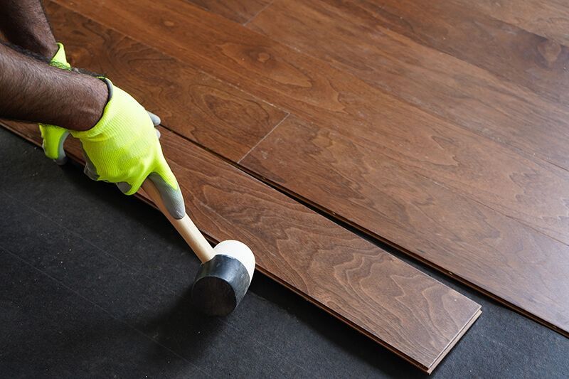 Person installing brown wood flooring, using a hammer and wearing yellow gloves.