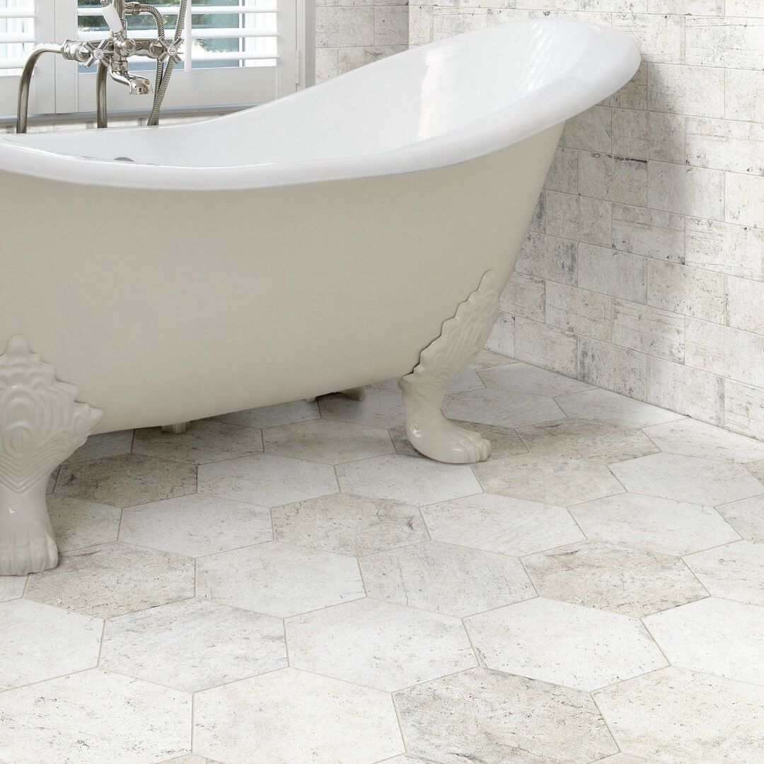 Clawfoot bathtub in a bathroom with hexagon-tiled floor and brick-like wall.
