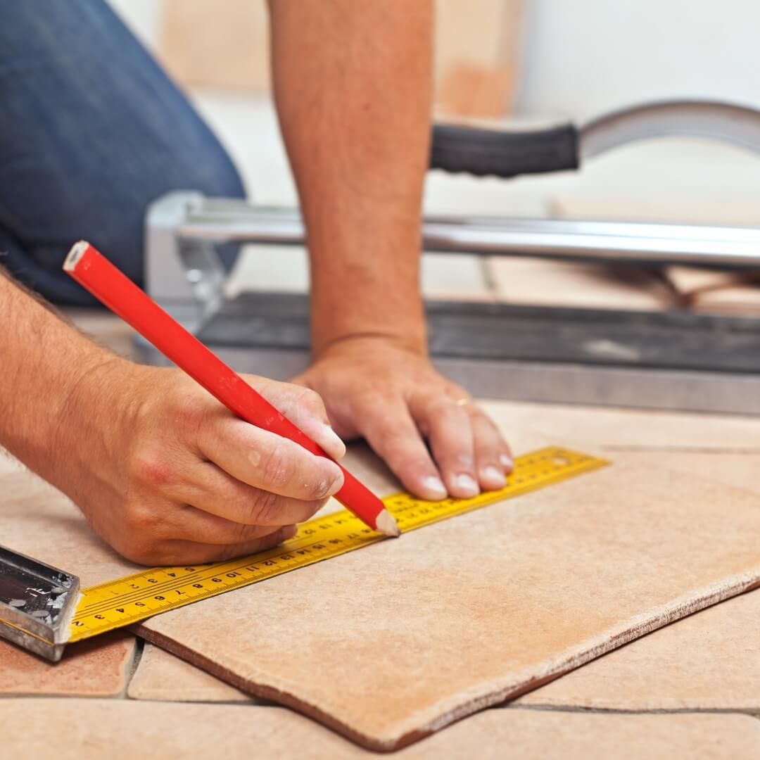 Person using a ruler and pencil to measure and mark a tan tile, with a tile cutter in the background.
