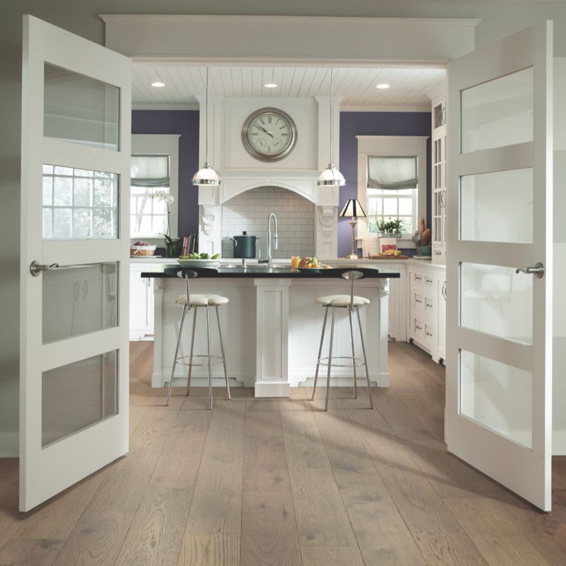Open white doors lead to a white kitchen with an island, stools, and a large clock.
