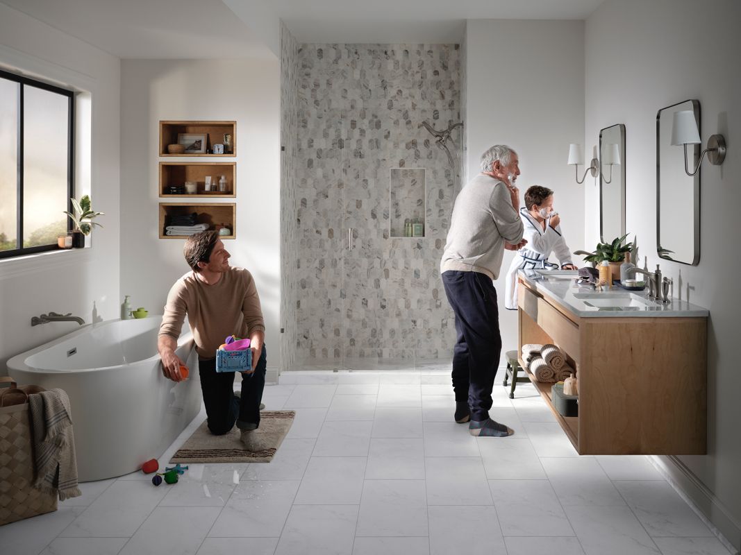 Family in a bright bathroom; one cleaning, two at the vanity. White tile, wood accents, and a large window.