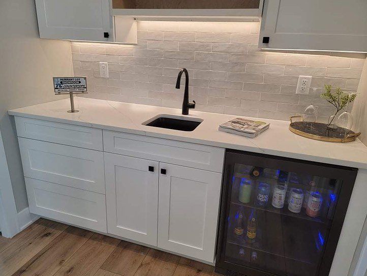 A white wet bar with black sink, faucet, and mini fridge filled with drinks, and a light stone backsplash.