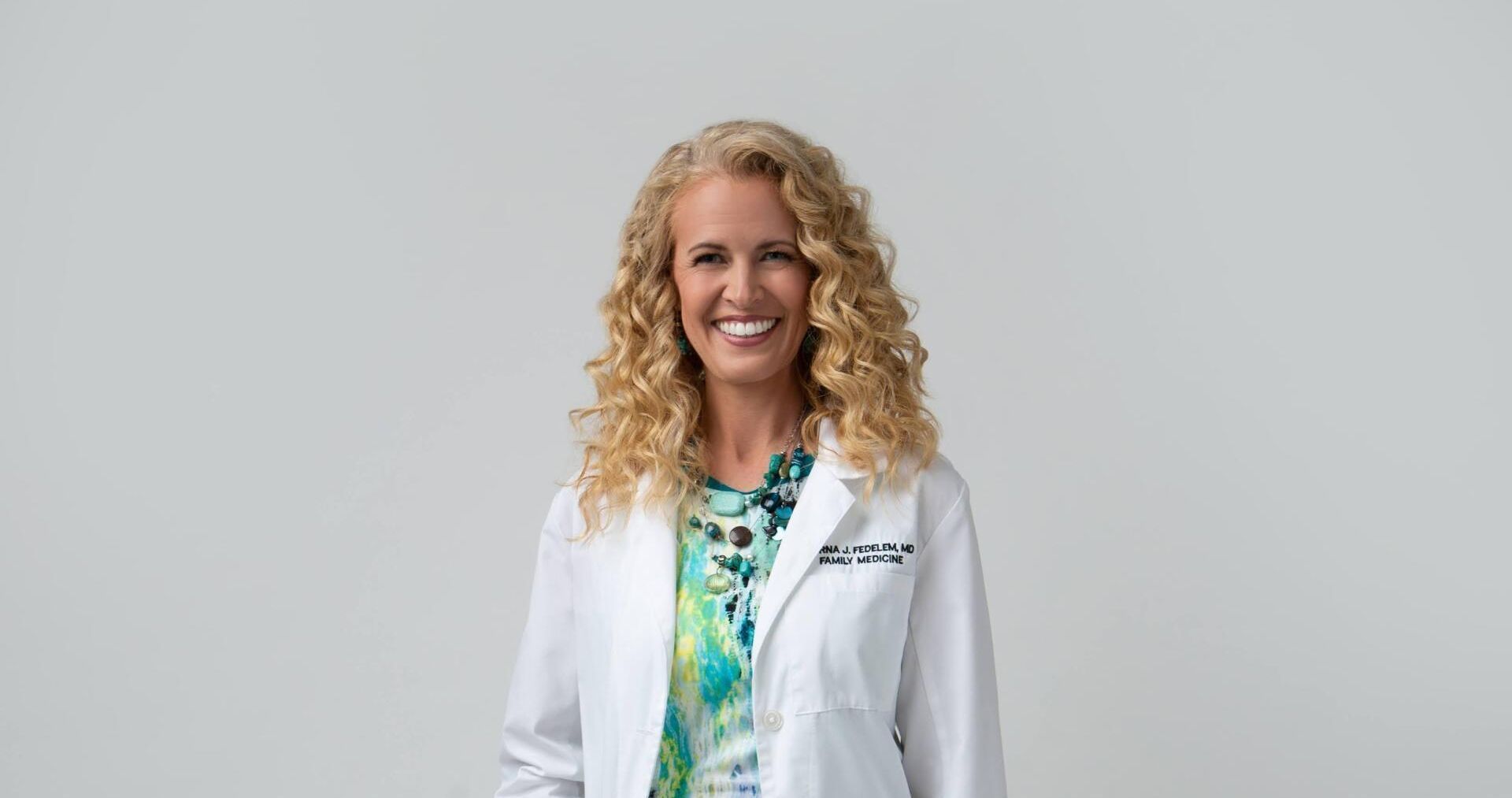 A woman is wearing a white lab coat and smiling.