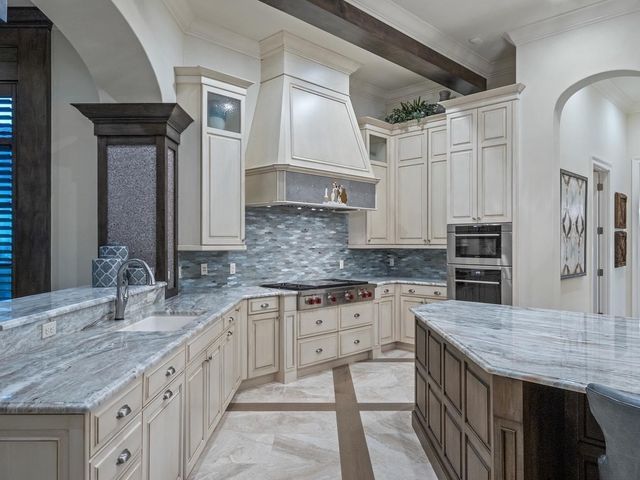 Elegant white kitchen with marble countertops, blue-gray backsplash, and vaulted ceiling beams