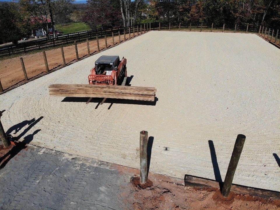 A small skid steer tractor leveling a sand arena with a wooden beam attachment. Outdoors, daylight.