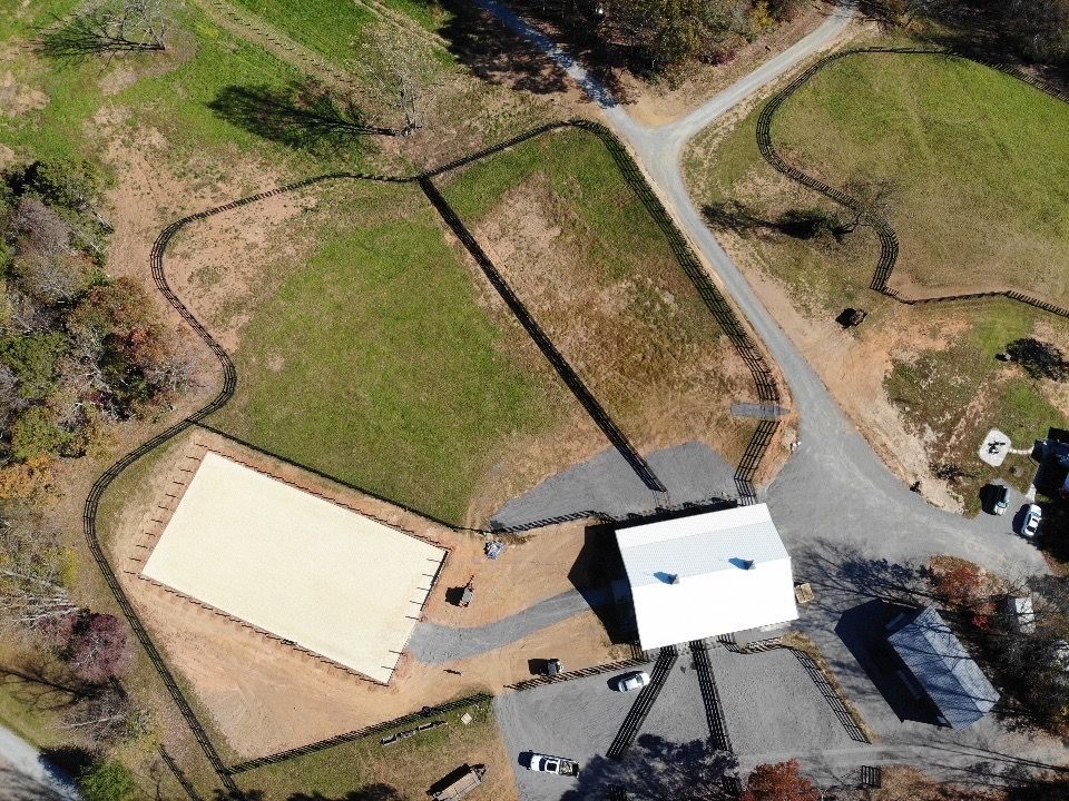 Aerial view of a white barn and outdoor riding arena surrounded by fields and a gravel parking lot.