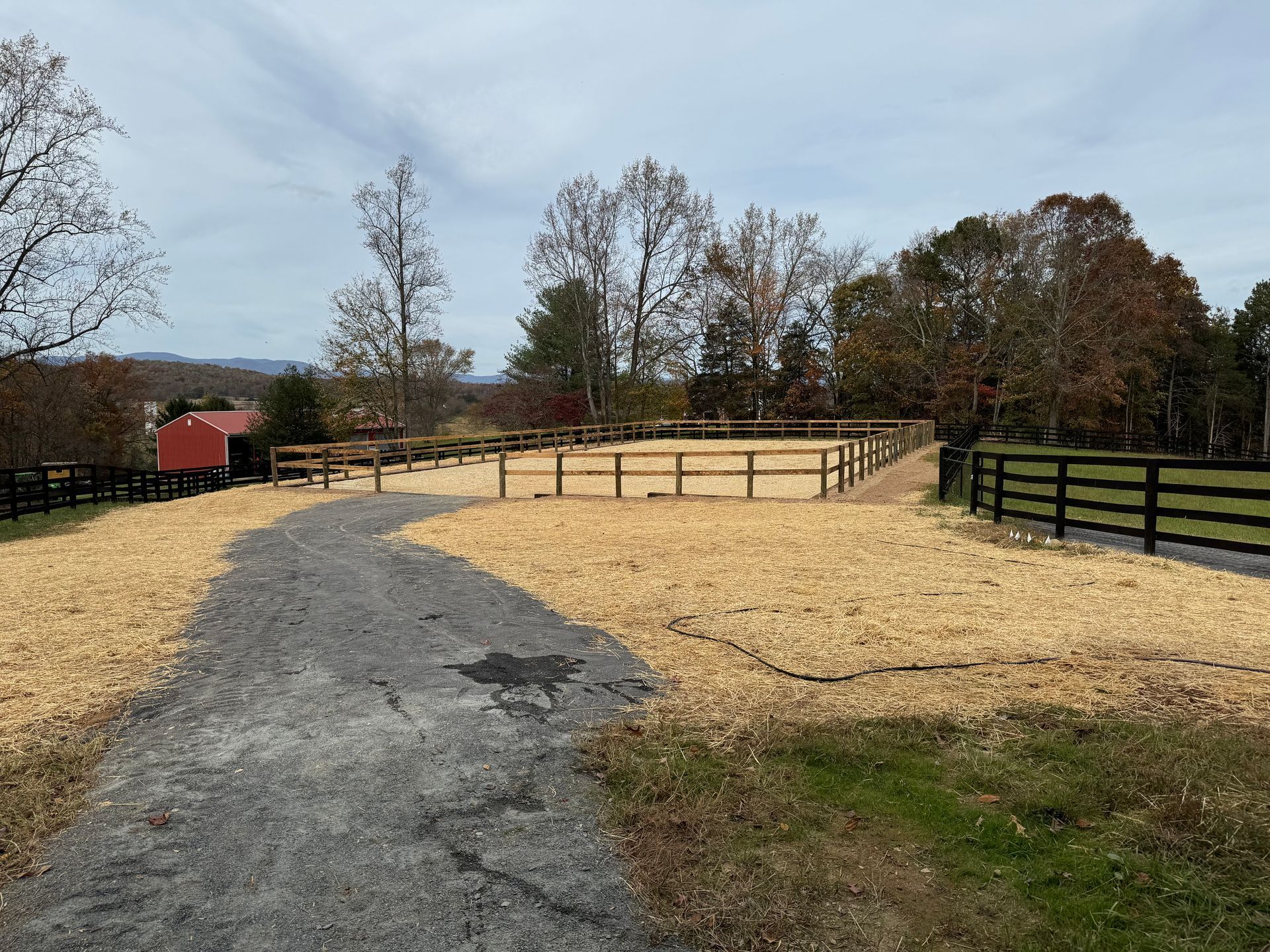Gravel driveway leads to a fenced riding arena filled with tan material; trees and red building in the background.