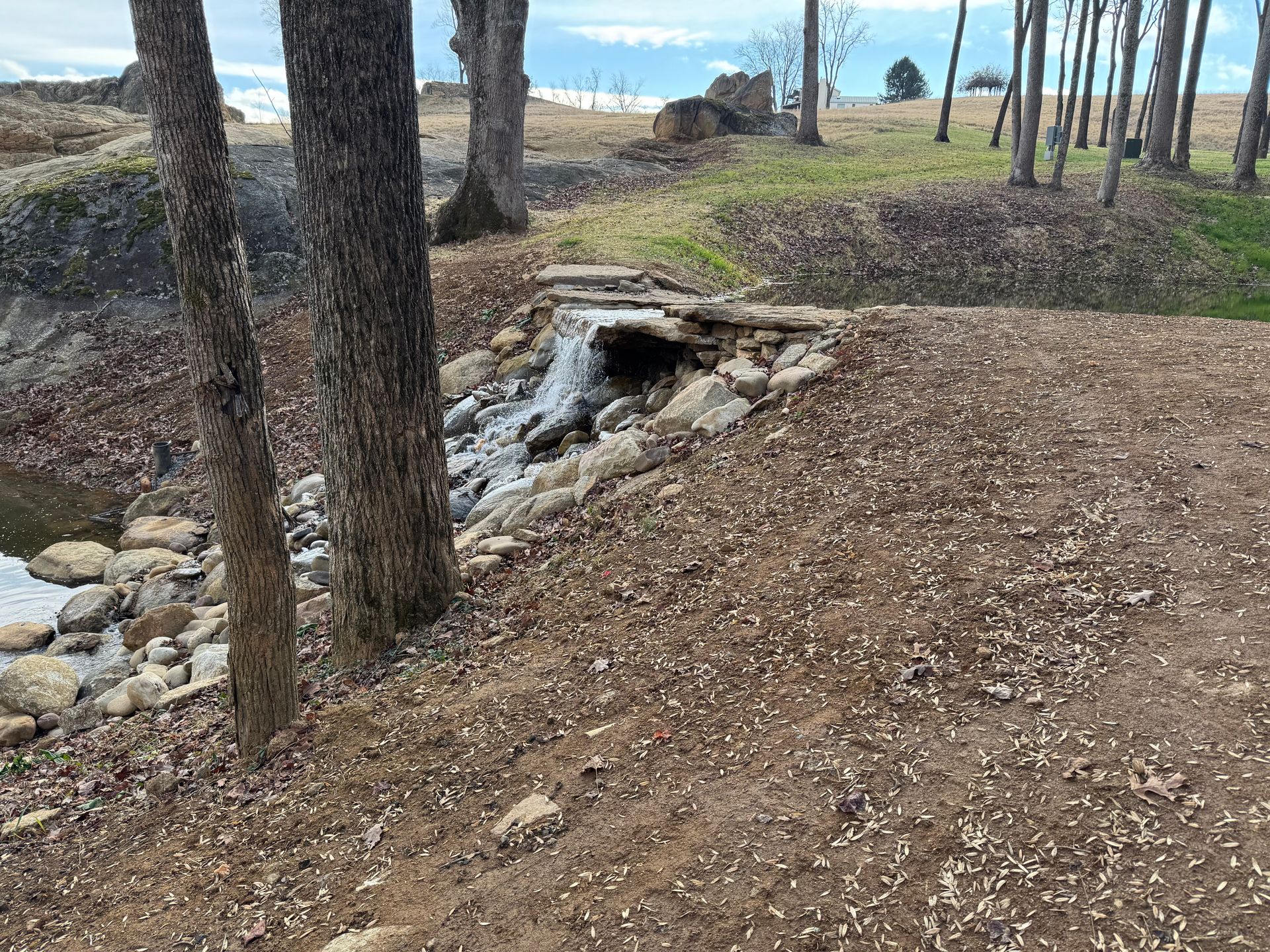 Small waterfall cascading under a stone bridge in a park. Brown leaves and trees surround.