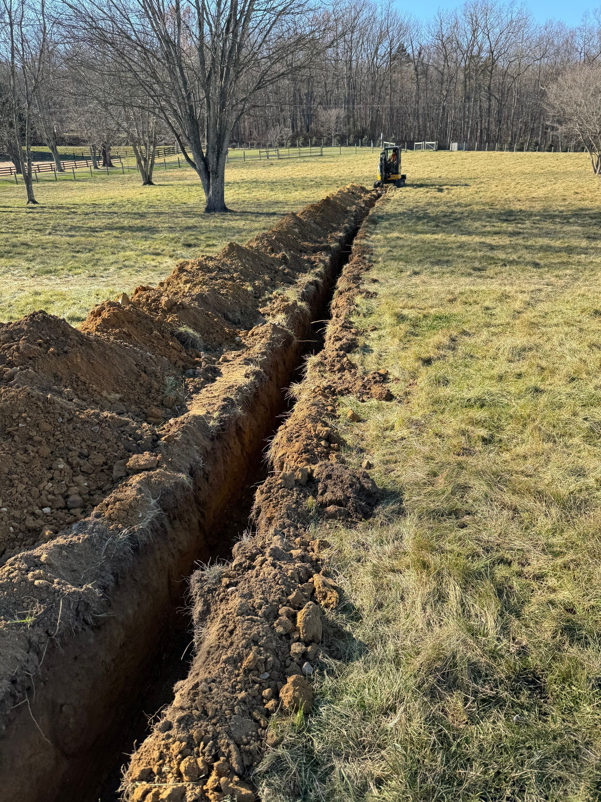 A long trench dug through a grassy field with a small excavator in the background.