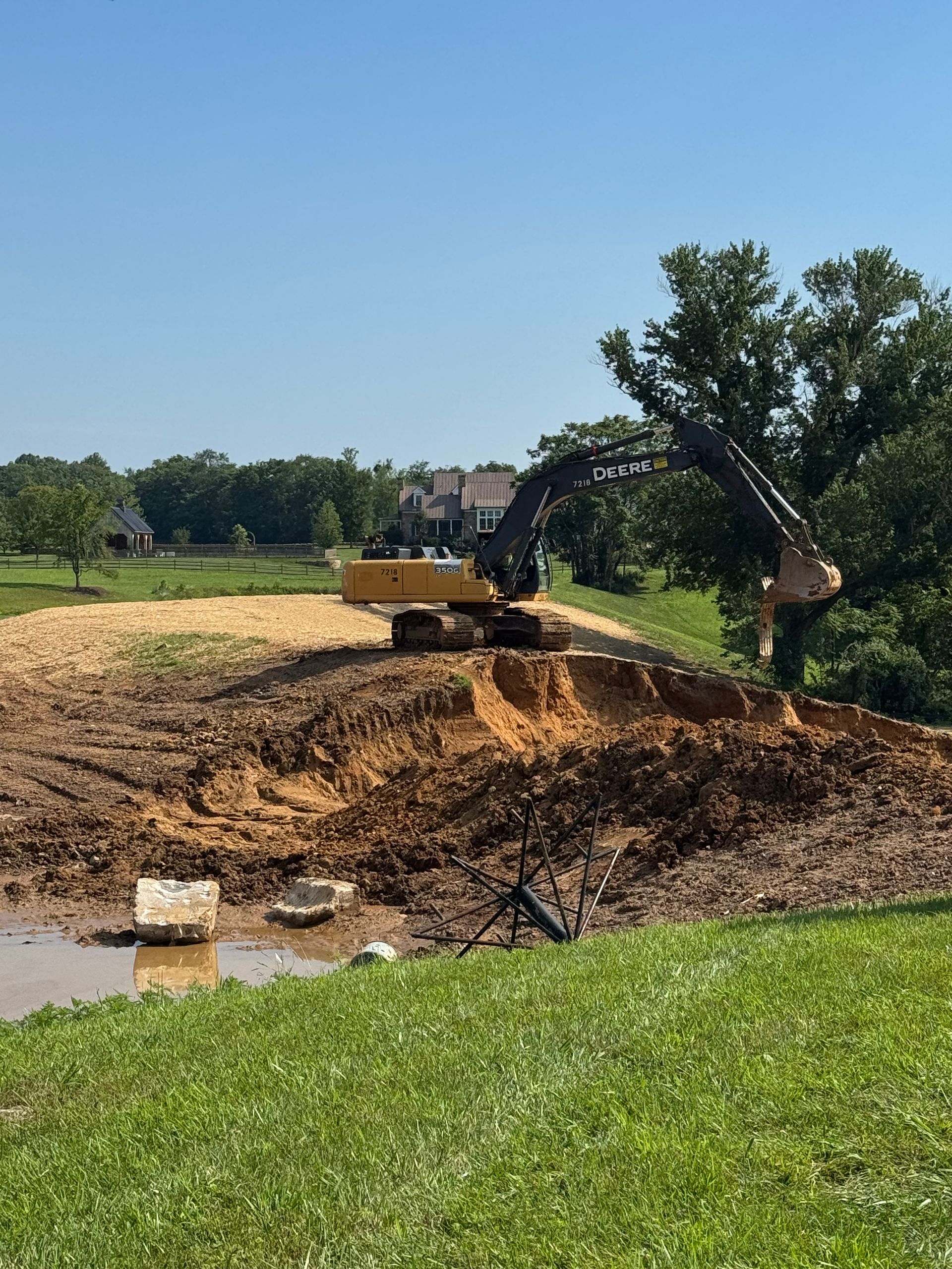 Yellow excavator working on a muddy hillside, near water. Houses and trees in the background under a blue sky.