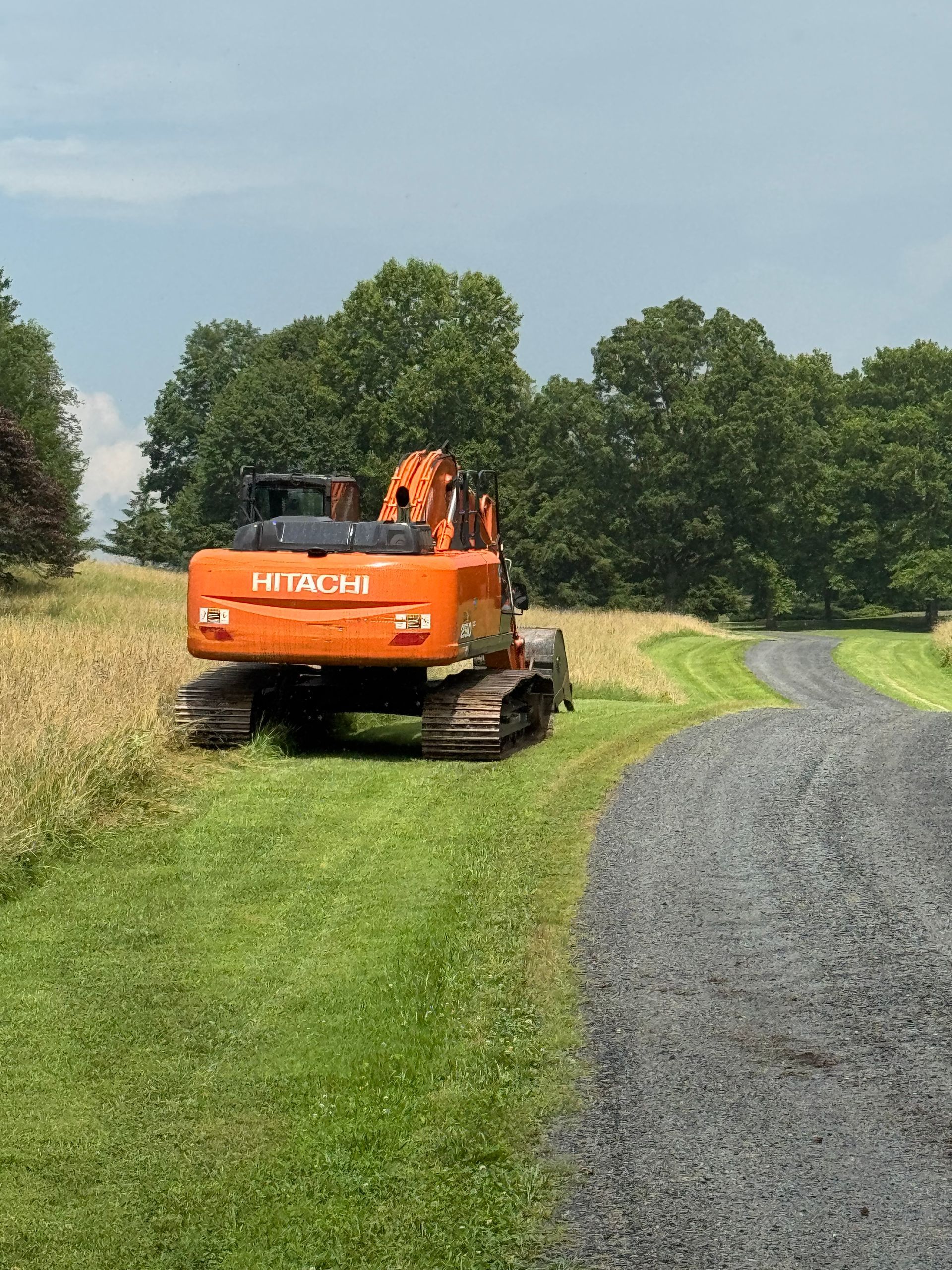 Orange Hitachi excavator parked beside a gravel driveway in a grassy field. Trees in the background.