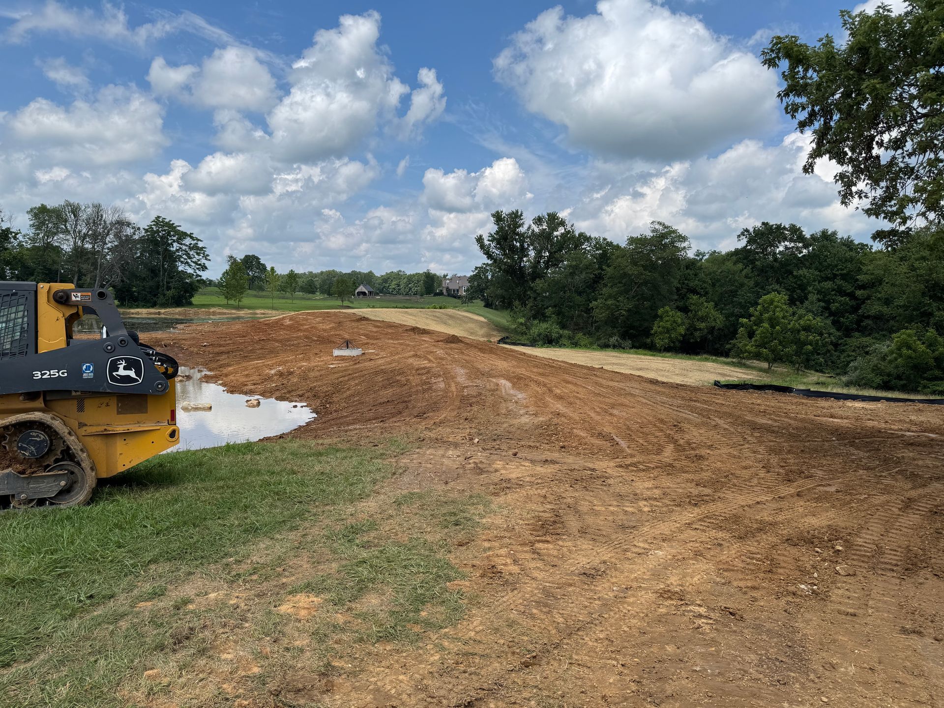 A John Deere skid steer next to a muddy construction site, trees and cloudy sky in background.