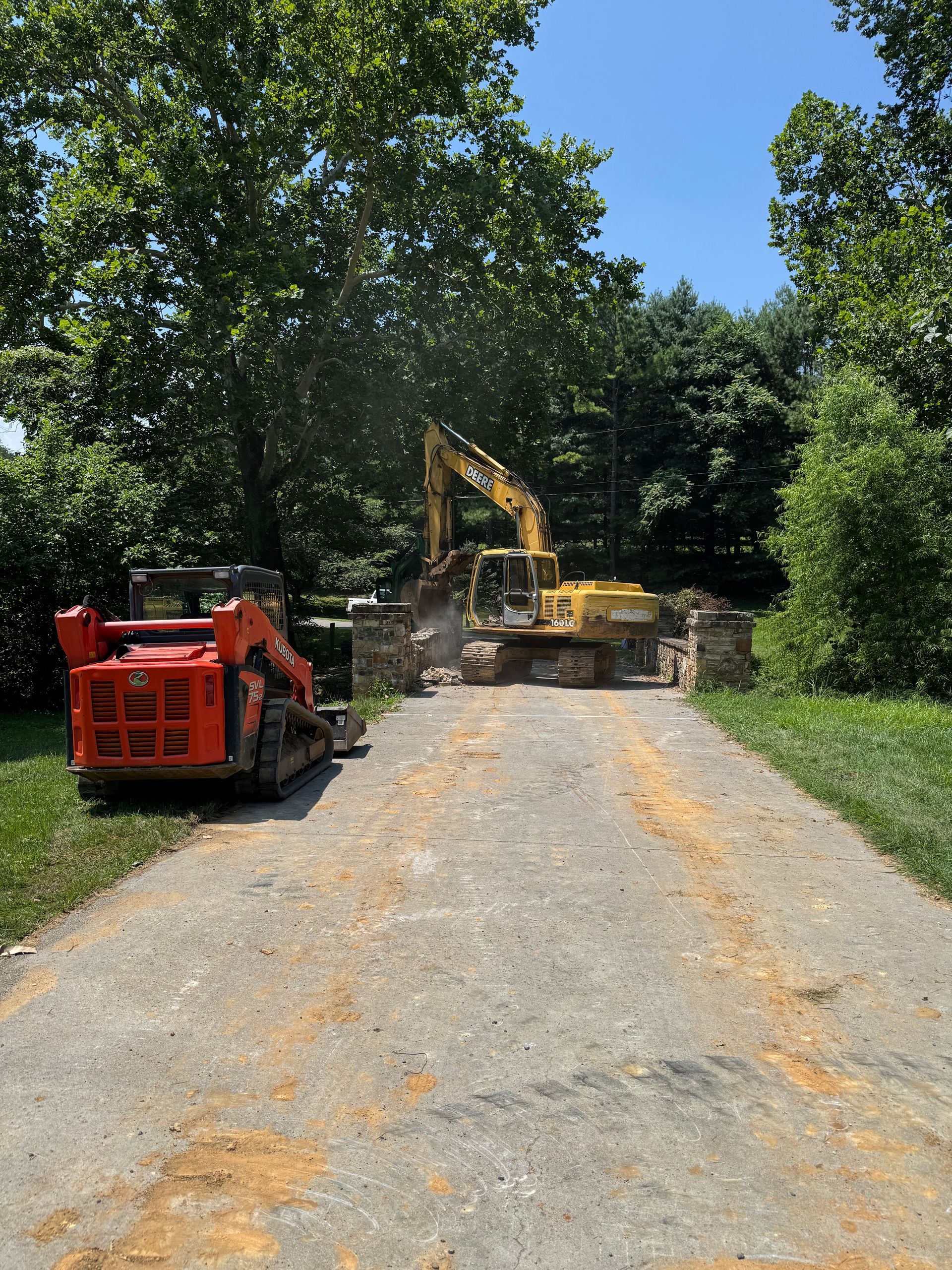 Construction equipment demolishing a stone structure on a gravel driveway, trees in the background, sunny day.