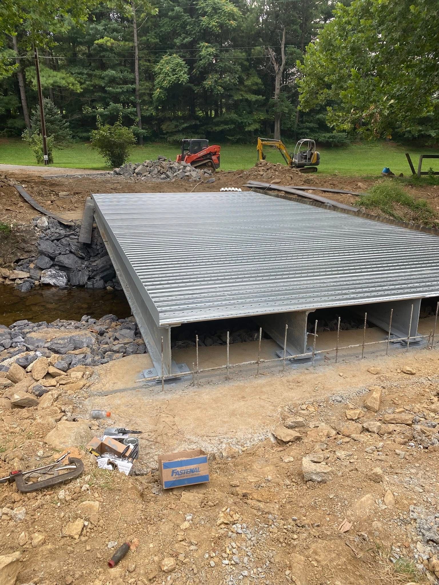 Metal bridge construction over a small stream. Dirt surrounds the bridge with machinery in the background.