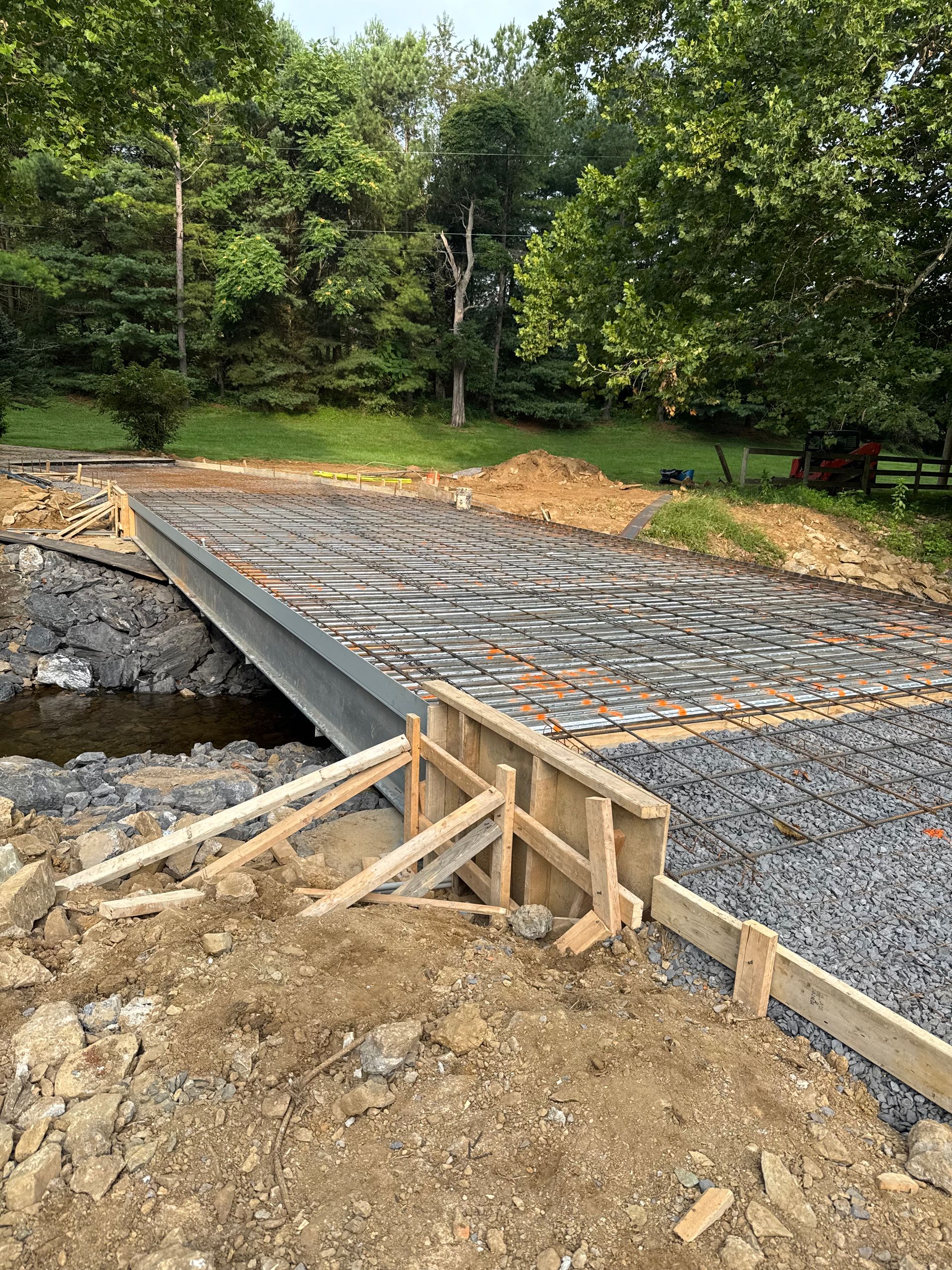 Bridge construction site. Steel beam spans stream, concrete rebar laid. Wooden forms in foreground. Trees in background.
