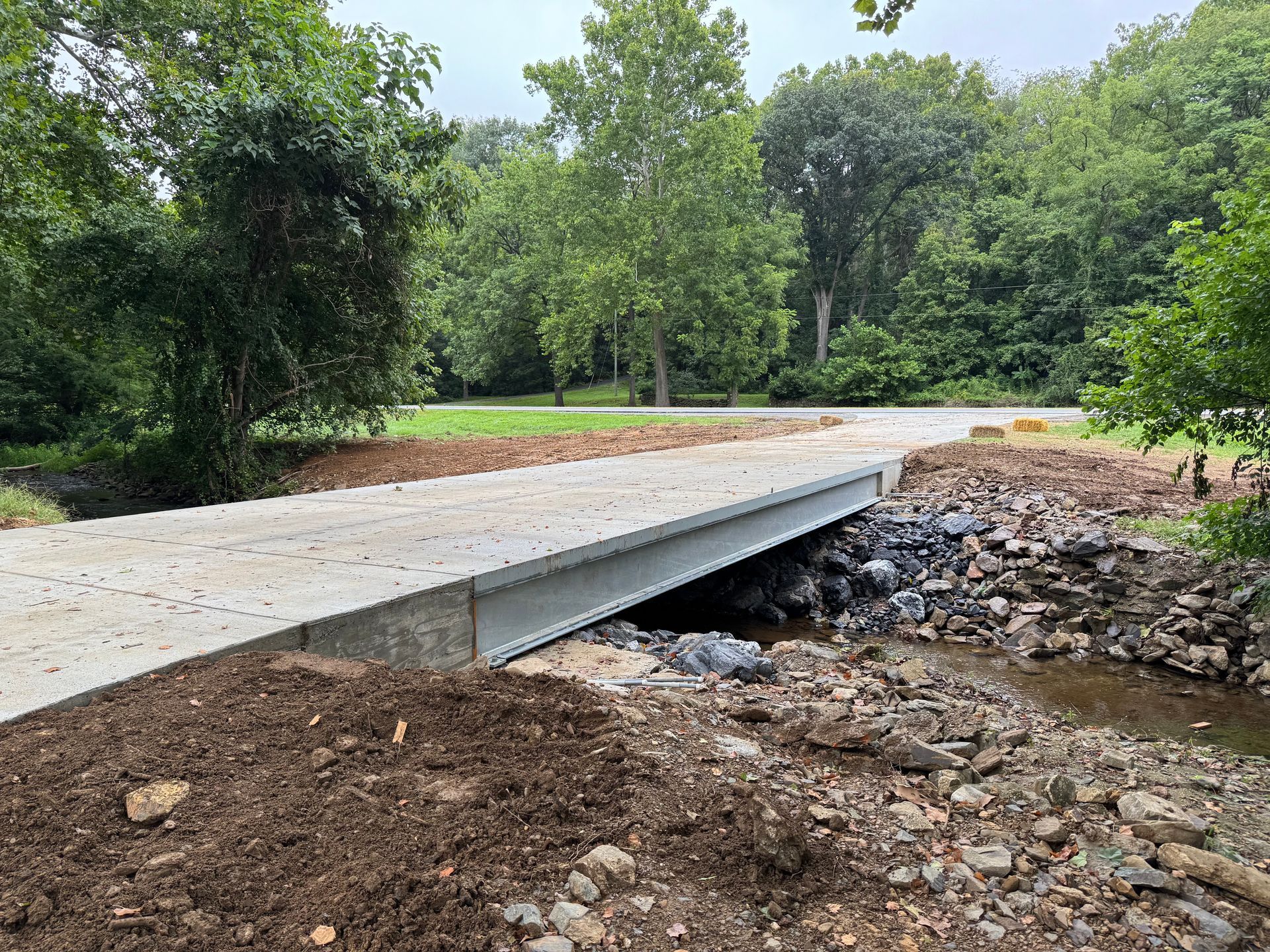 Concrete bridge over a creek in a park setting. Brown dirt and rocks visible. Green trees in the background.