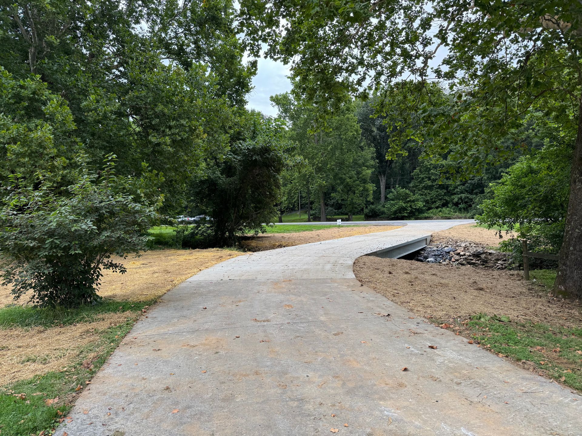 Gravel driveway curves through a park-like setting with trees and landscaping on a cloudy day.
