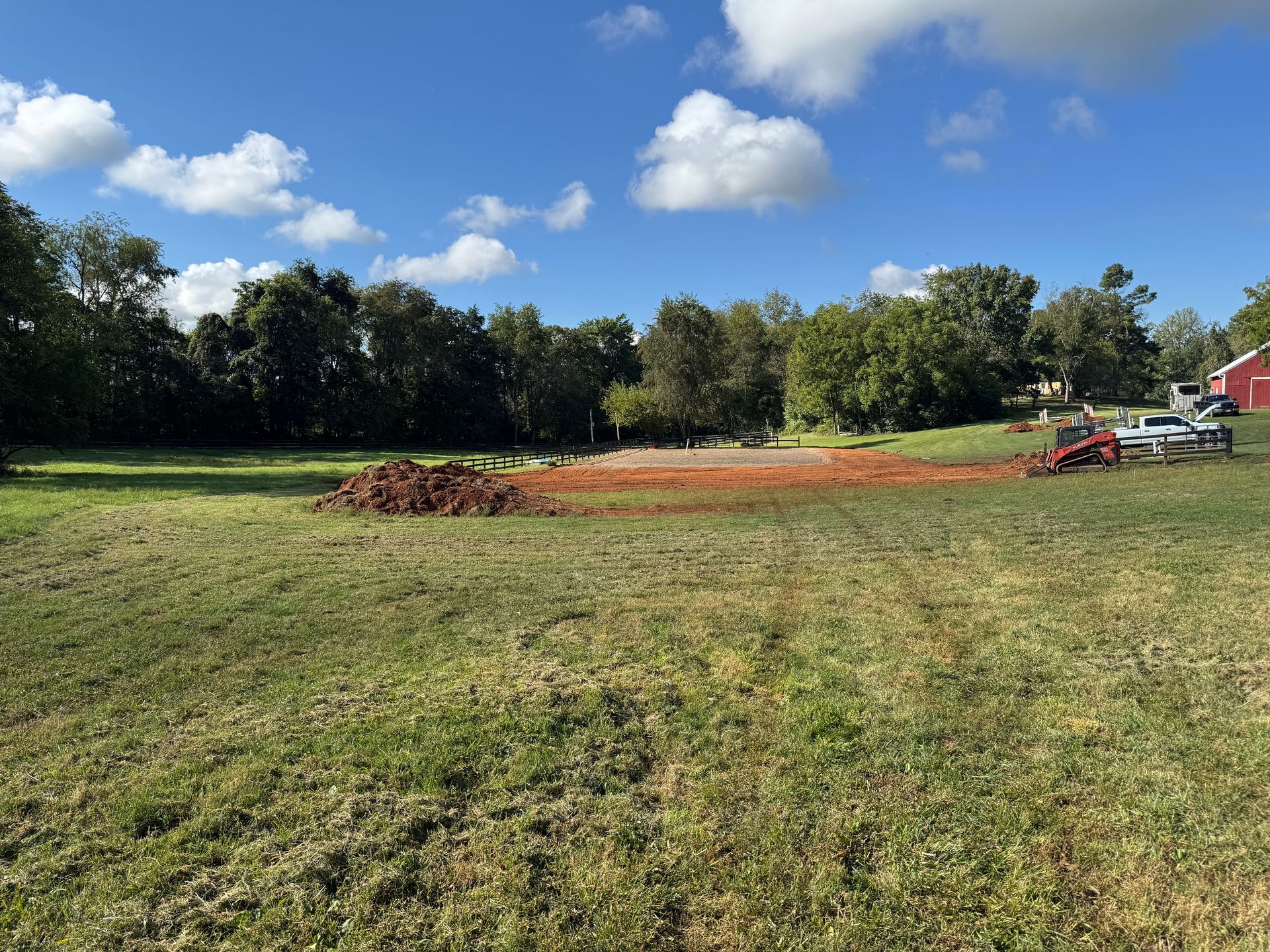 Grassy field with a cleared area of reddish-brown soil, trees in the background, blue sky.