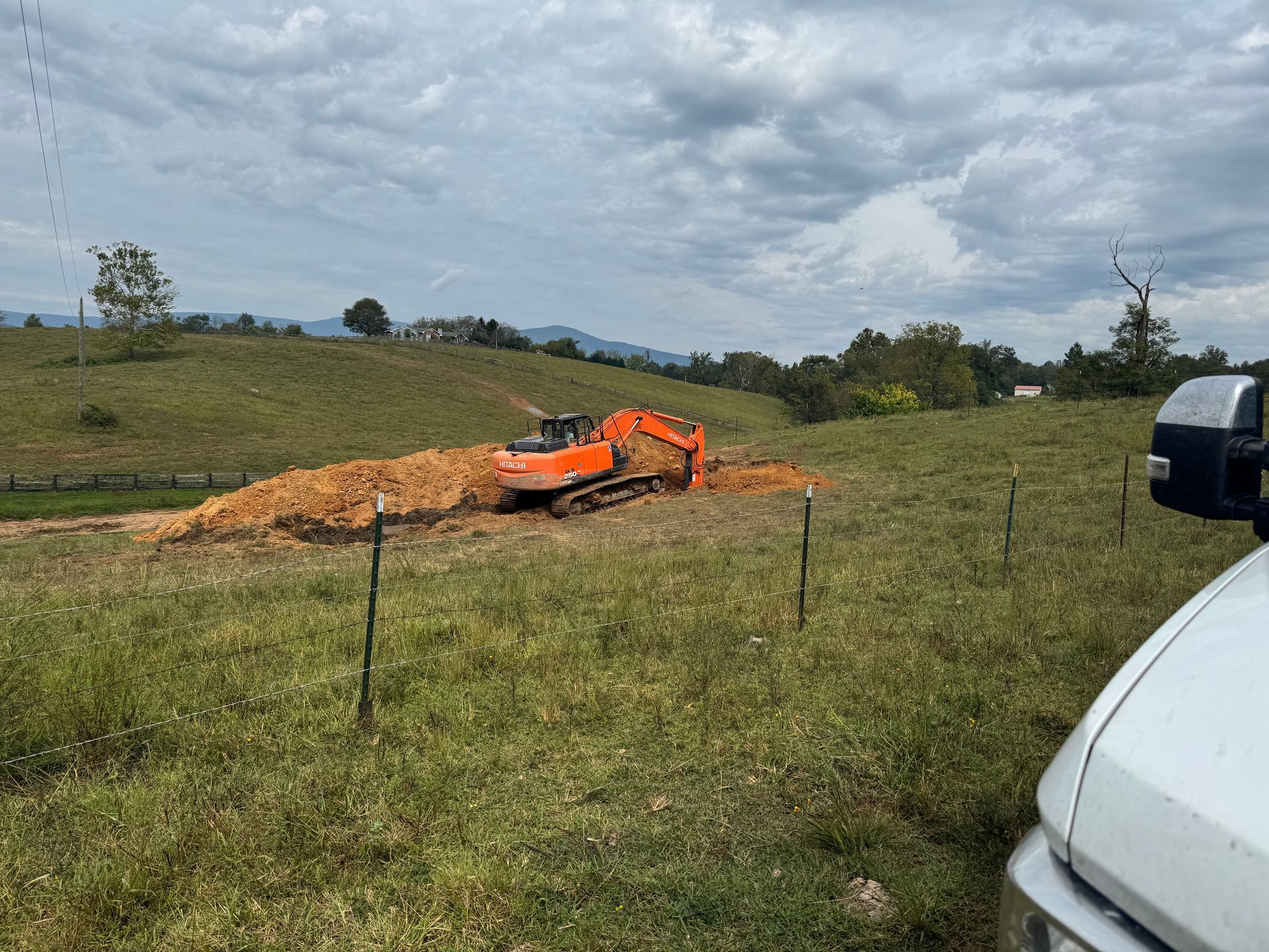 An orange excavator digs in a grassy field. Hills and a cloudy sky are in the background.
