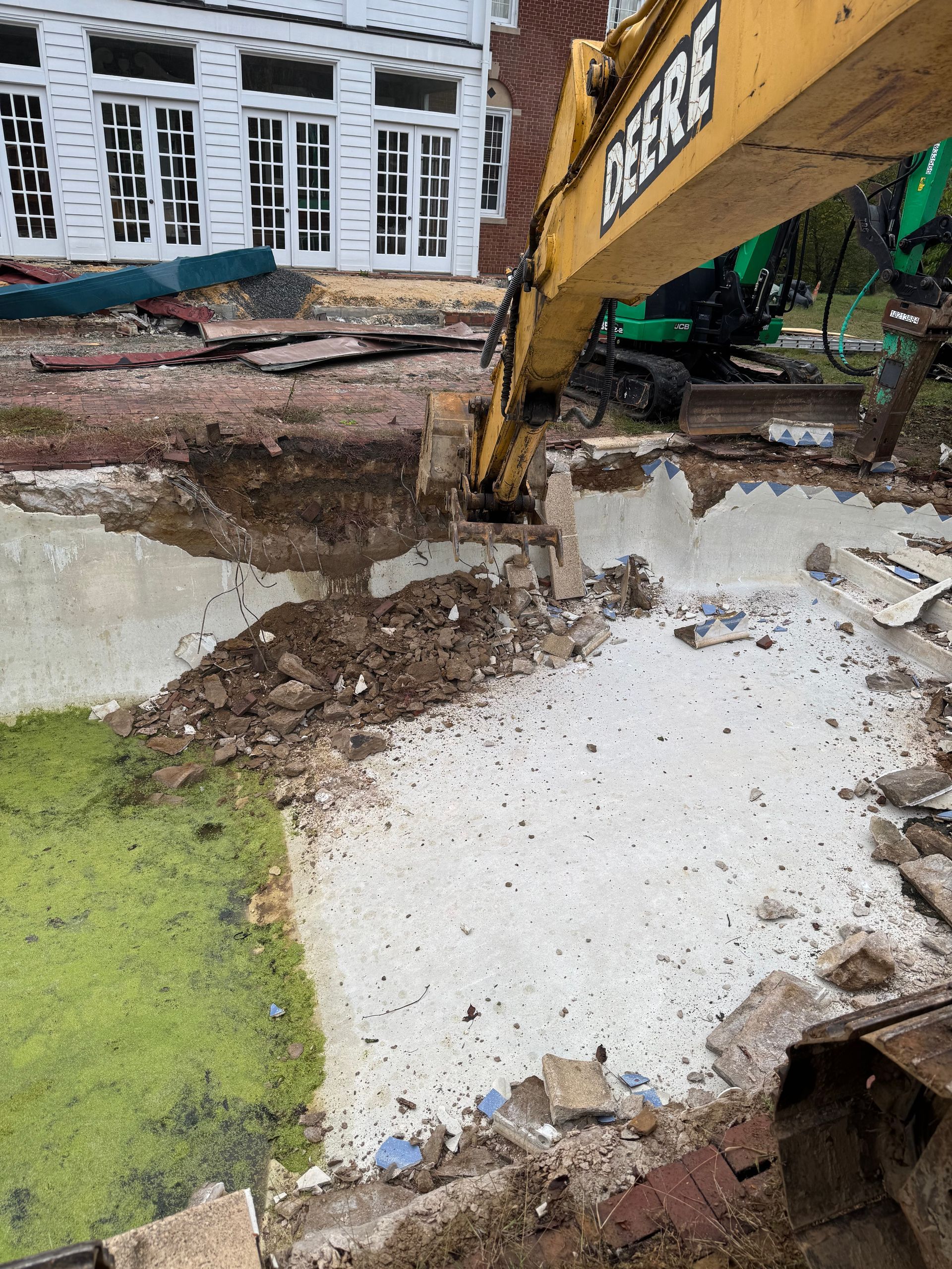 An excavator demolishing a concrete structure near a building; debris and water present.