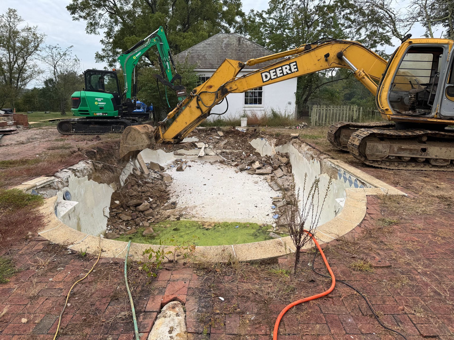 Two excavators demolish a pool in a yard. One is yellow, the other green.