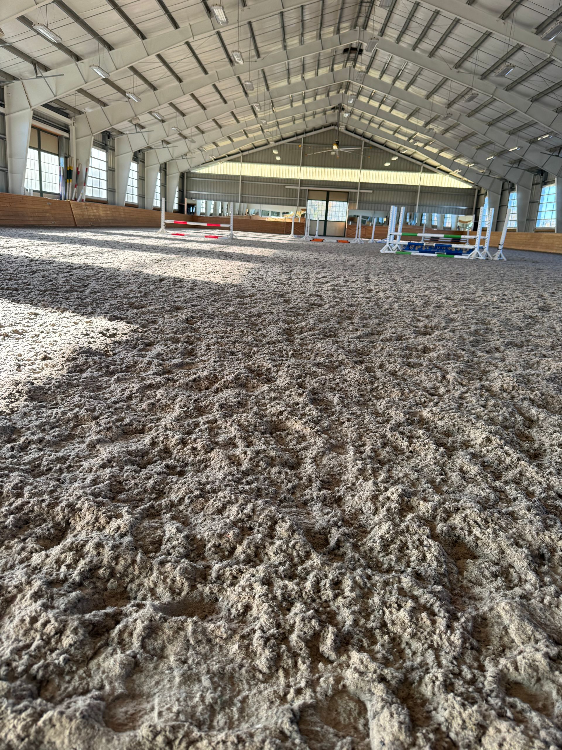 Indoor equestrian arena with tan footing, jumps, and open ceiling.